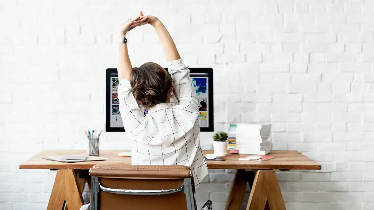 Une femme s'étire sur sa chaise de bureau devant son ordinateur, illustrant l'importance d'une bonne posture pour le travail prolongé.