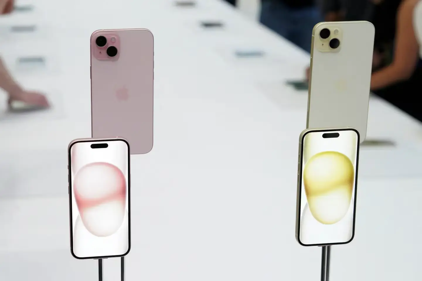 Deux iPhone 15 Plus, l'un rose et l'autre jaune, exposés sur un présentoir, avec leur écran allumé montrant des fonds d'écran colorés.