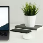 Bureau minimaliste avec un ordinateur portable affichant le message "Work Hard Anywhere", accompagné d'un smartphone, d'une souris et d'une plante verte en pot sur un fond blanc.