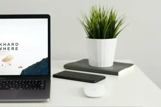 Bureau minimaliste avec un ordinateur portable affichant le message "Work Hard Anywhere", accompagné d'un smartphone, d'une souris et d'une plante verte en pot sur un fond blanc.