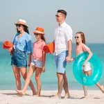 Une famille marchant sur une plage sous un ciel dégagé, avec en arrière-plan une personne sur une bouée flottante.