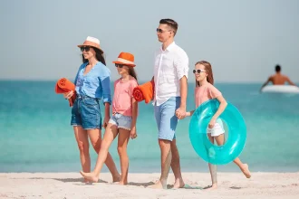 Une famille marchant sur une plage sous un ciel dégagé, avec en arrière-plan une personne sur une bouée flottante.