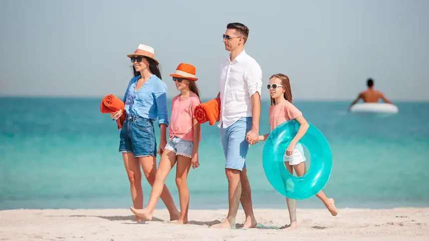 Une famille marchant sur une plage sous un ciel dégagé, avec en arrière-plan une personne sur une bouée flottante.