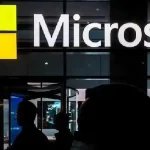 Logo de Microsoft sur une façade vitrée, avec des silhouettes de personnes au premier plan.